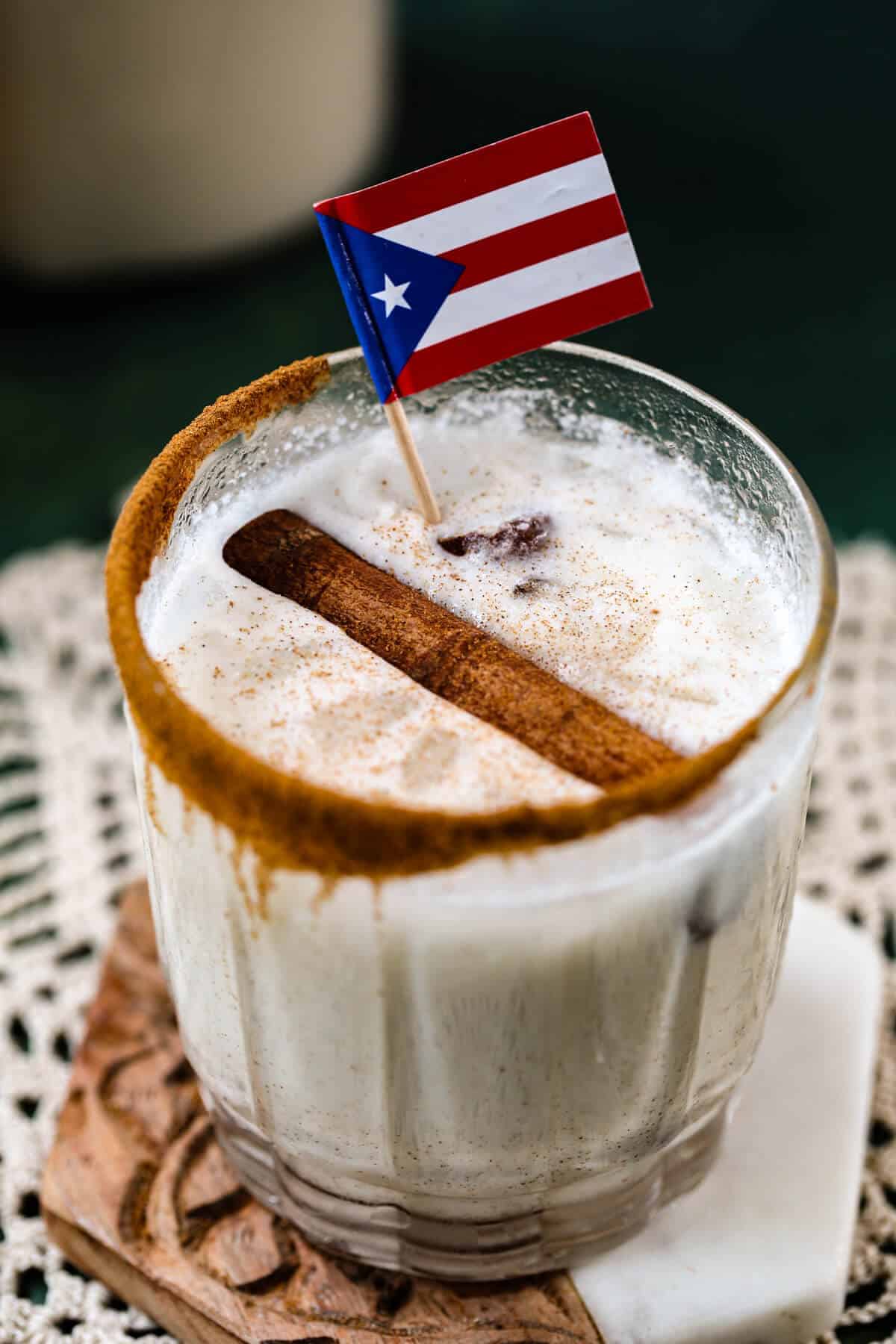 Glass of classic Puerto Rican coquito with a cinnamon-sugar rim, garnished with a cinnamon stick and Puerto Rican flag toothpick, dusted with ground cinnamon, served on a white lace doily.