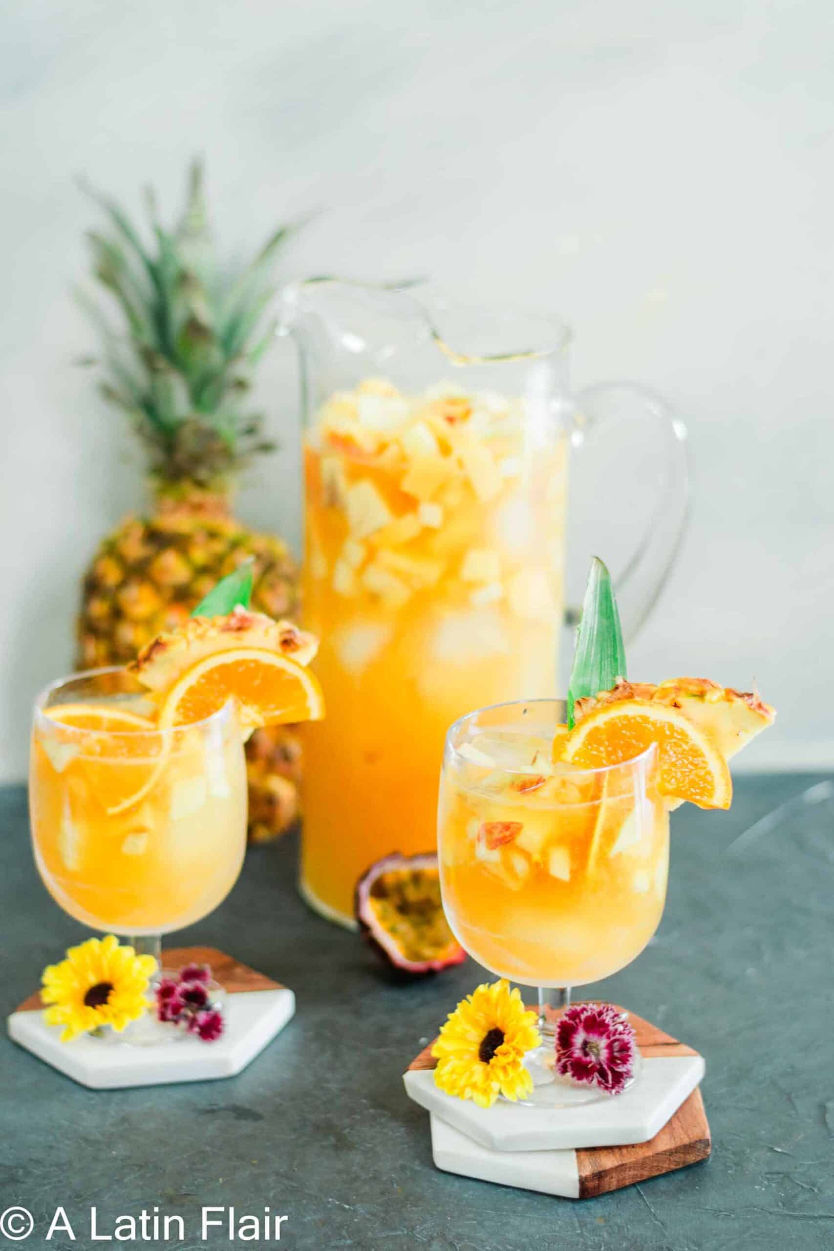 Two glasses of Puerto Rican sangria garnished with orange slices, pineapple wedges, and pineapple leaves, served alongside a pitcher of tropical sangria with fresh fruit, decorated with yellow and pink flowers on white marble coasters.