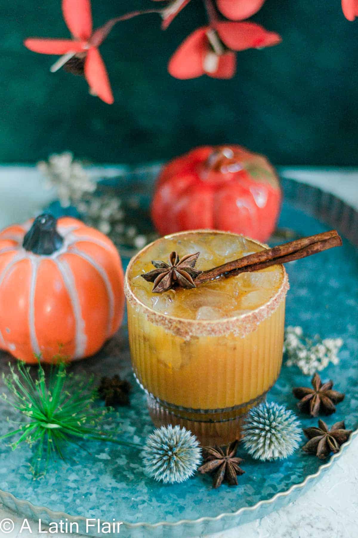 A pumpkin spice margarita in a textured glass with a sugar-rimmed edge, garnished with a star anise, cinnamon stick, and lime wheel, surrounded by decorative mini pumpkins and fall foliage on a turquoise tray.