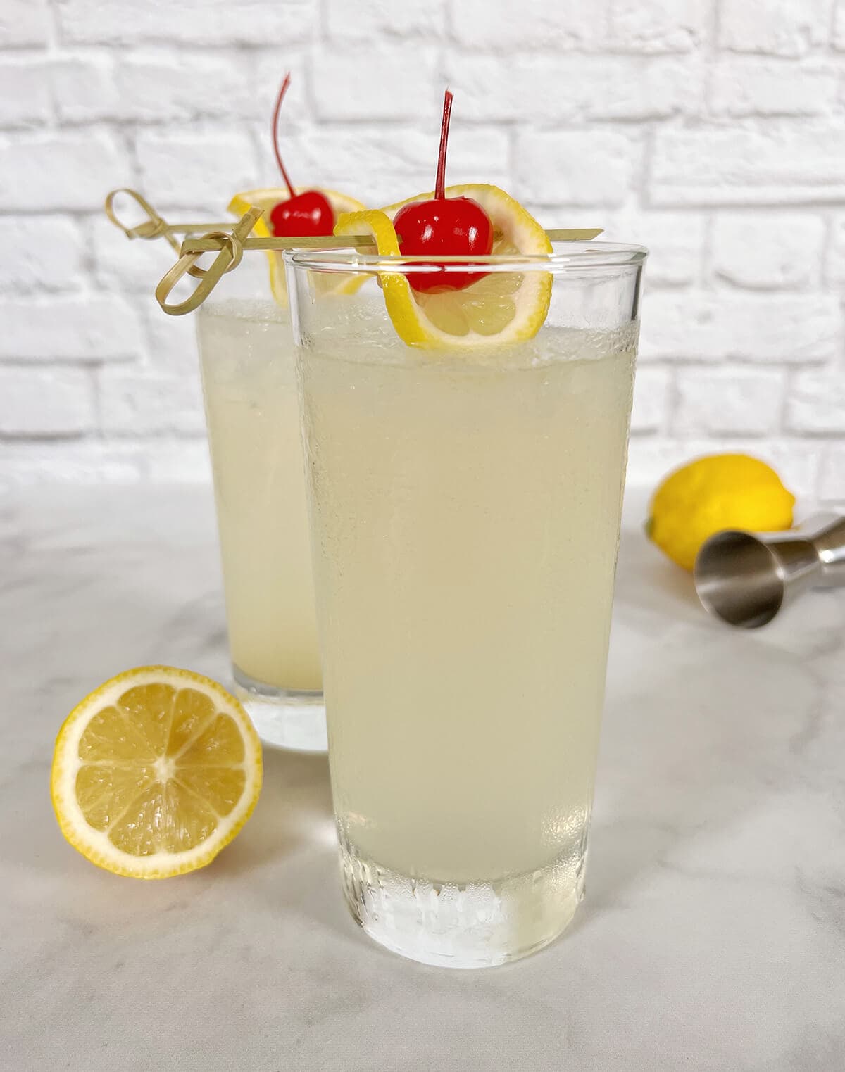 Two tall glasses of Rum Collins cocktail with pale lemon-colored liquid filled with ice, garnished with lemon twists, lemon slices, and maraschino cherries on cocktail picks, displayed on a white marble surface with fresh lemons and a jigger against a white brick wall background.