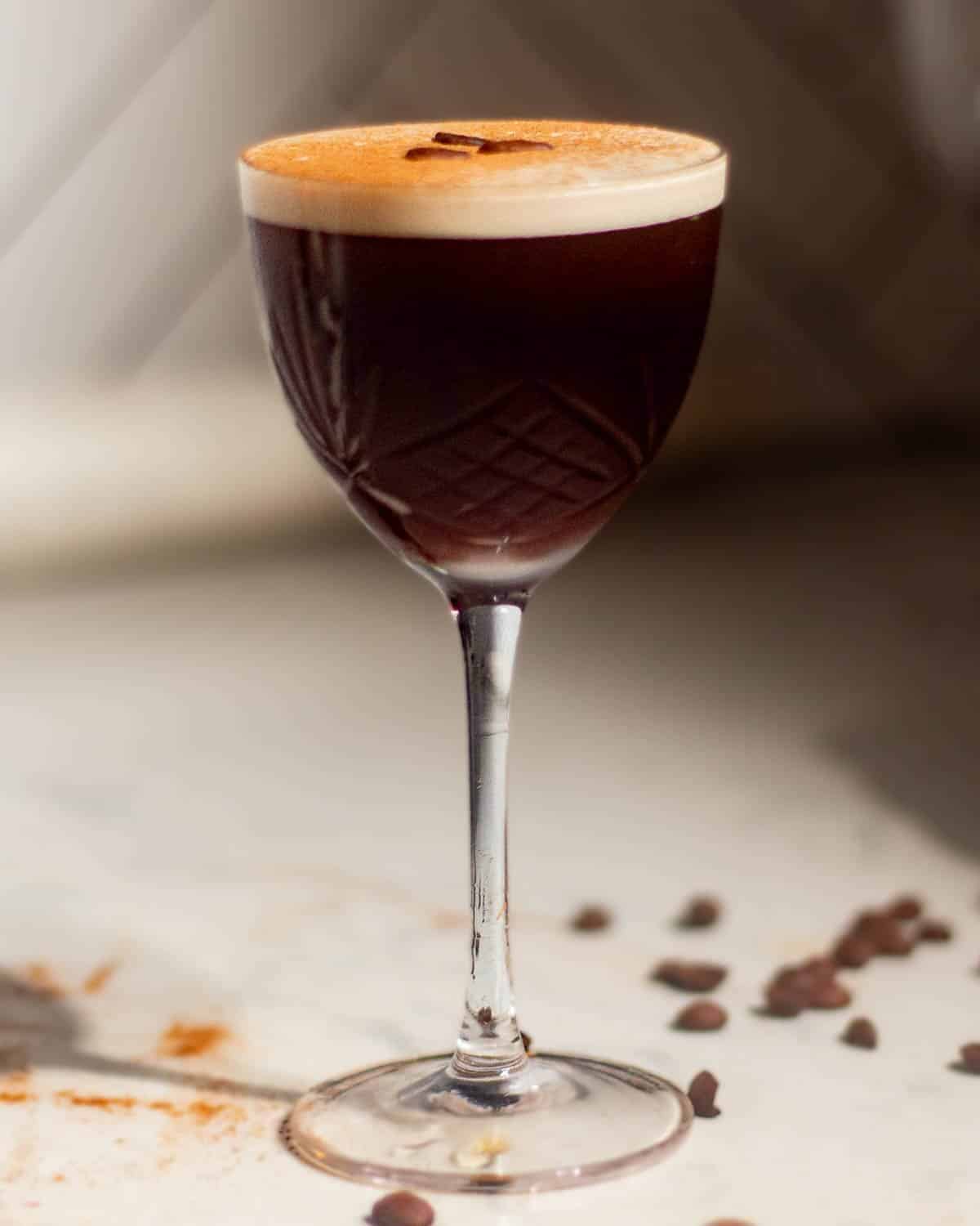 Rum espresso martini in a textured coupe glass with a thick creamy foam top garnished with three coffee beans, displayed on a white surface with scattered coffee beans against a neutral background.