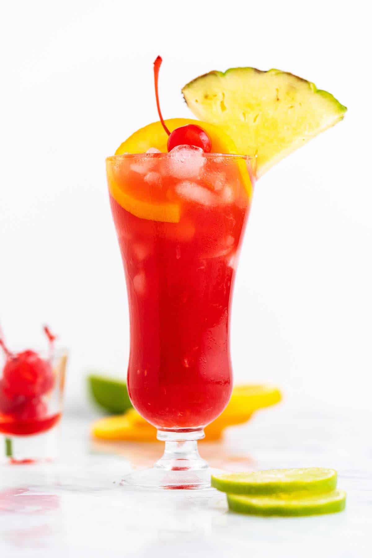 Frozen rum runner cocktail in a tall hurricane glass with layered red and orange frozen liquid, garnished with a large pineapple wedge, orange slice, and maraschino cherry, displayed on a white surface with lime slices and tropical fruit in the background.