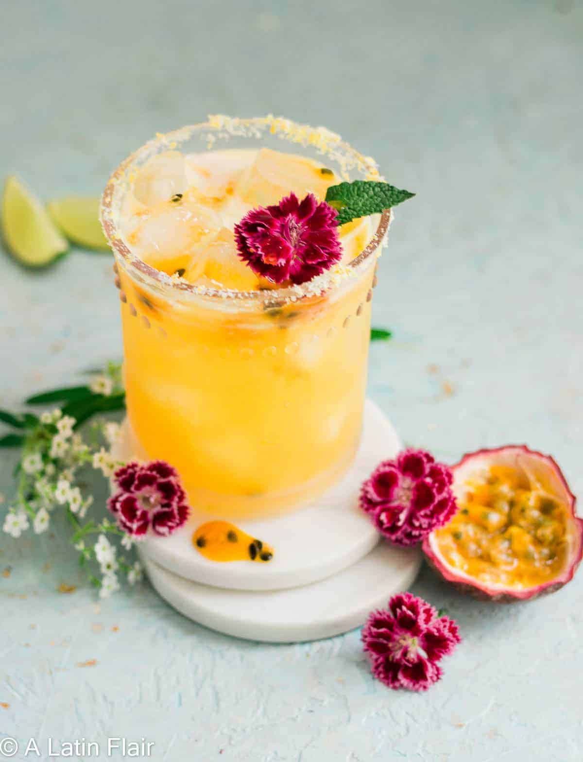 Skinny passion fruit margarita recipe in a salt-rimmed glass garnished with edible flowers and fresh mint, served with passion fruit halves and lime wedges.