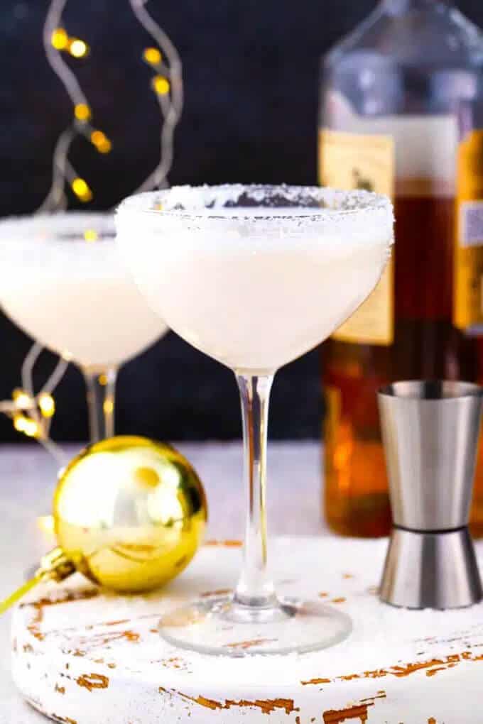 Two creamy white snowflake martinis in sugar-rimmed coupe glasses on a distressed white tray, with a gold Christmas ornament, cocktail shaker, and twinkling lights in the background.