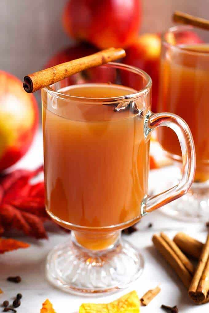 Spiced apple hot toddy in a clear glass mug with handle, garnished with a cinnamon stick, surrounded by fresh red apples, cinnamon sticks, autumn leaves, and whole cloves.