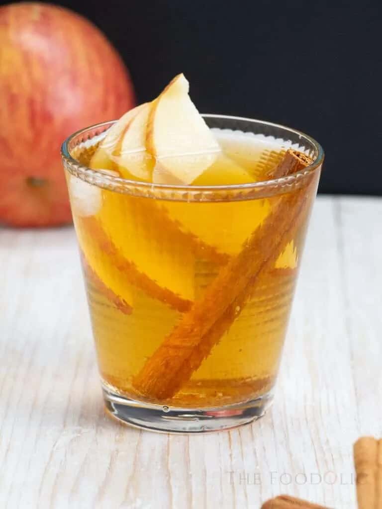 Glass of spiced apple spritz cocktail with golden amber liquid and ice, garnished with a fresh apple slice and cinnamon stick, served in a sugar-rimmed rocks glass on a white wooden surface with a fresh apple in the background against a dark backdrop.