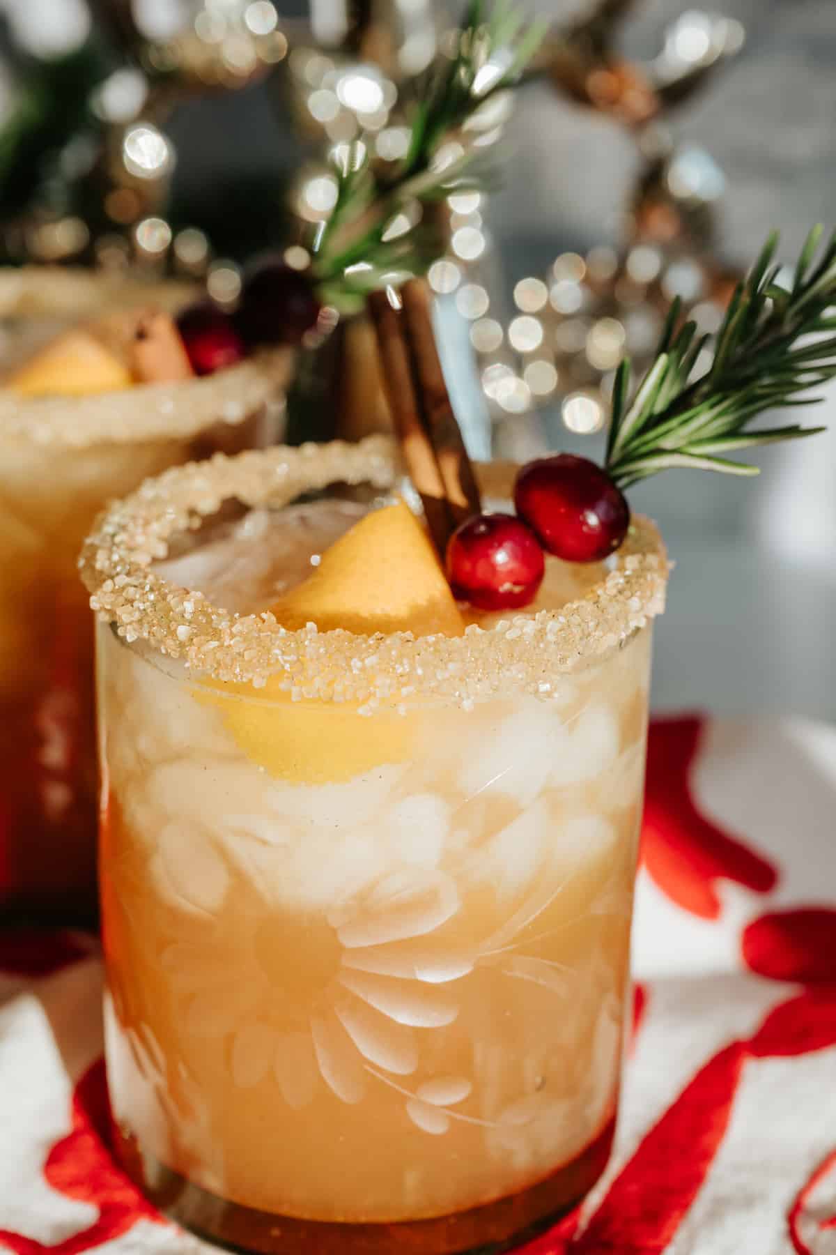 Spiced holiday margarita in a sugar-rimmed glass filled with ice, garnished with apple slices, fresh cranberries, cinnamon stick, and rosemary, with twinkling holiday lights in the background.