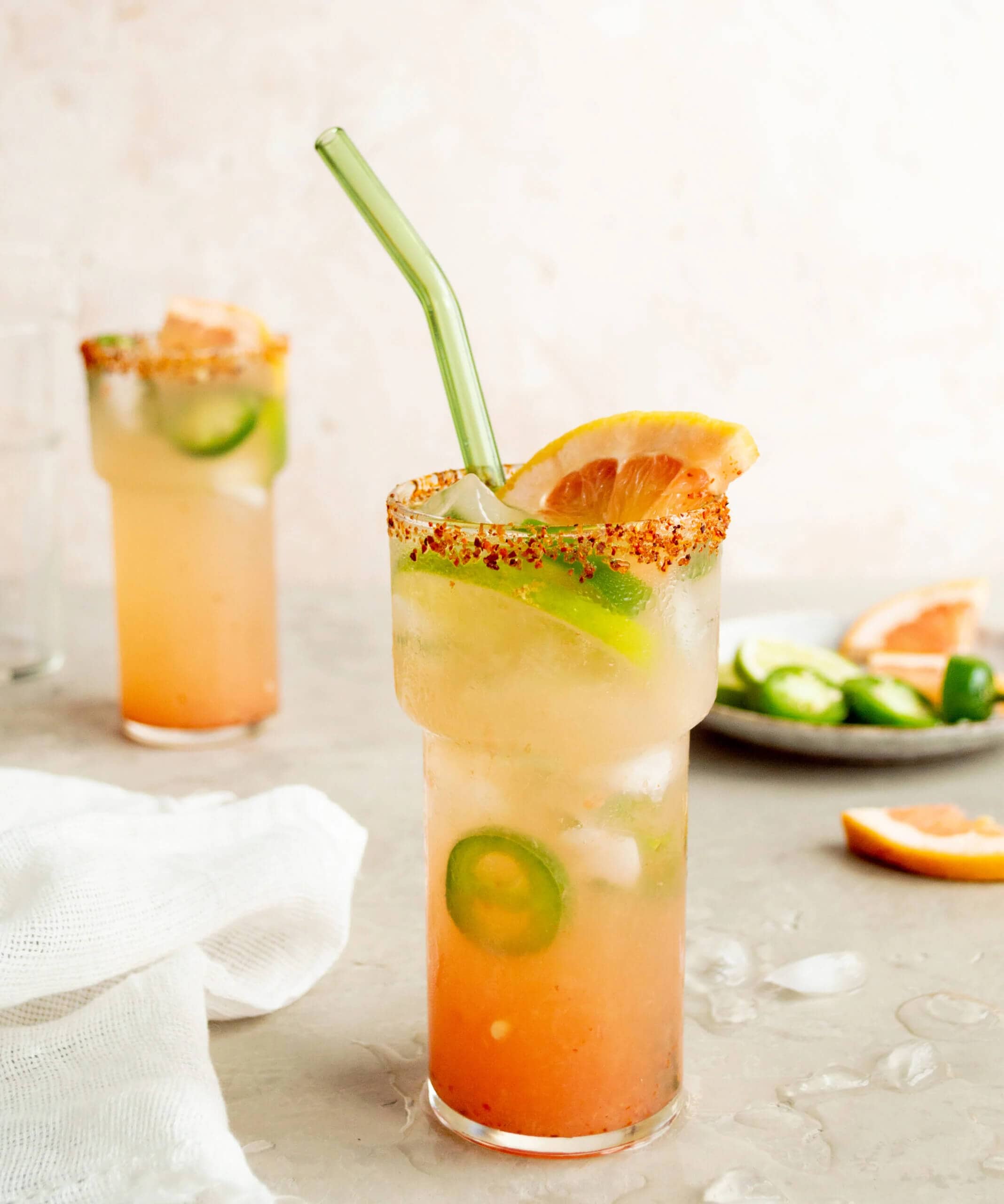 A spicy grapefruit ranch water cocktail in a tall glass with a chili lime salt rim, garnished with a grapefruit slice, celery stalk, and jalapeño slices, with a second cocktail and fresh jalapeños in the background.