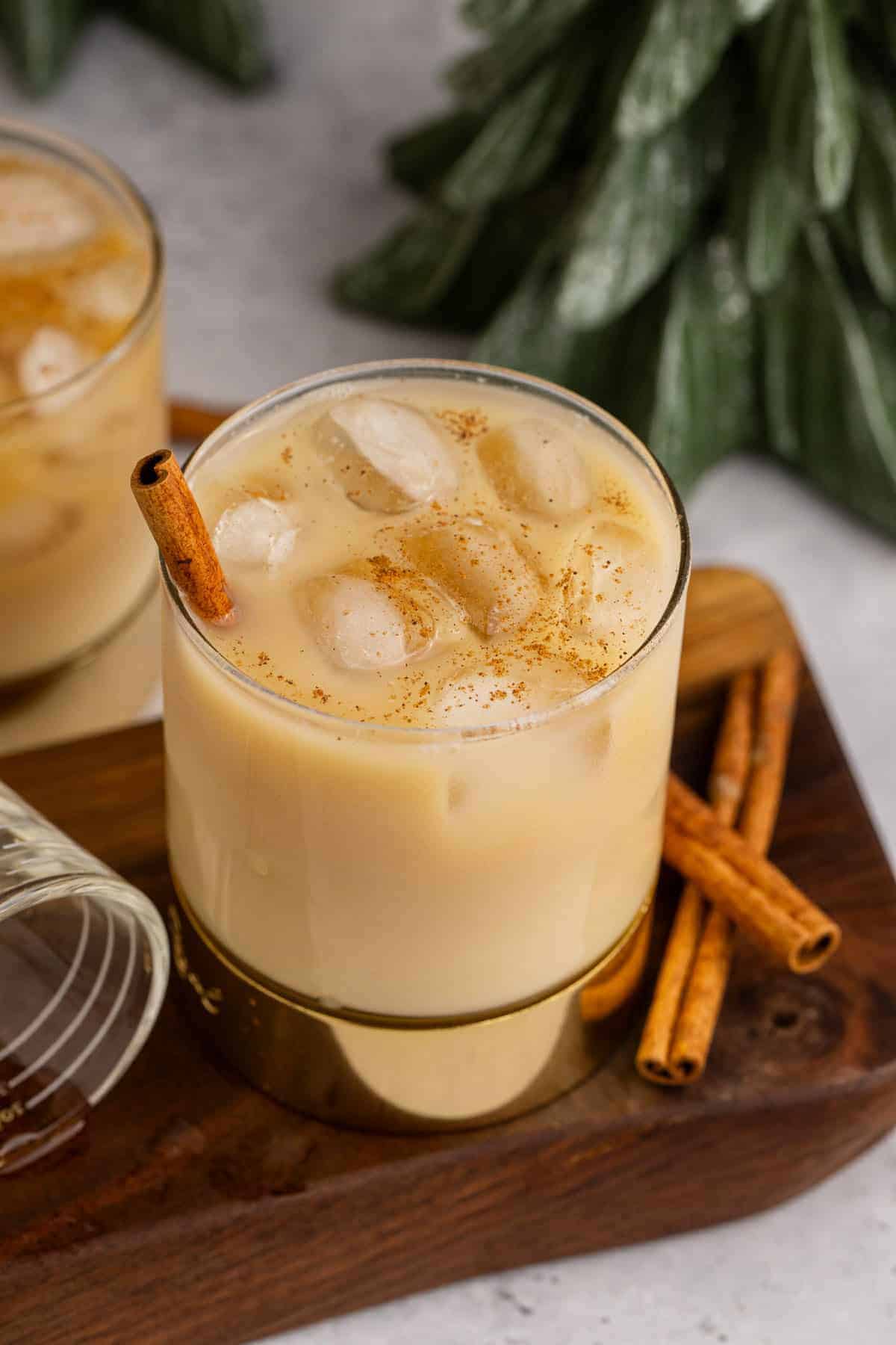 Glass of spiked rum eggnog with ice in a gold-rimmed glass, garnished with a cinnamon stick and dusted with ground nutmeg, served on a wooden board with extra cinnamon sticks and festive evergreen branches in the background.