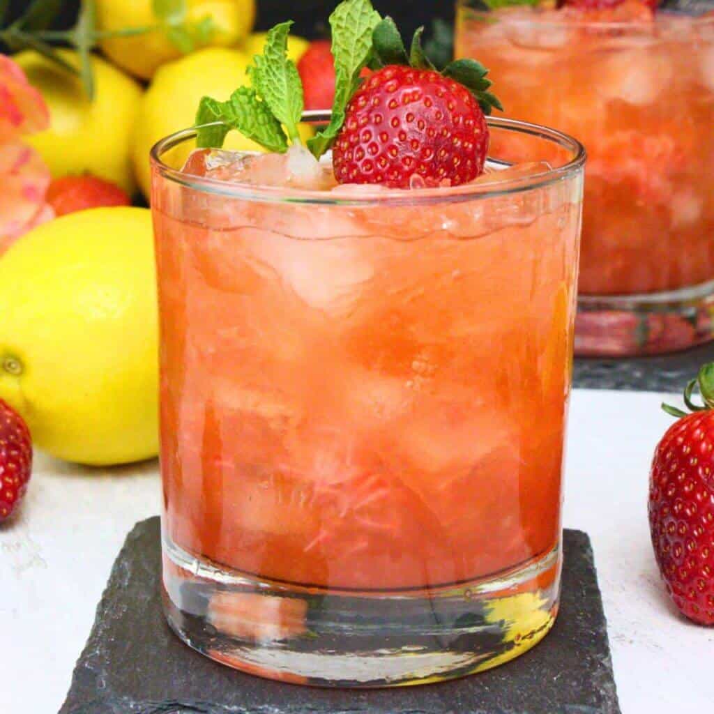 A strawberry tequila smash in a rocks glass with muddled strawberries visible throughout, garnished with a fresh strawberry and mint sprig, served on a slate coaster with fresh lemons and strawberries in the background.