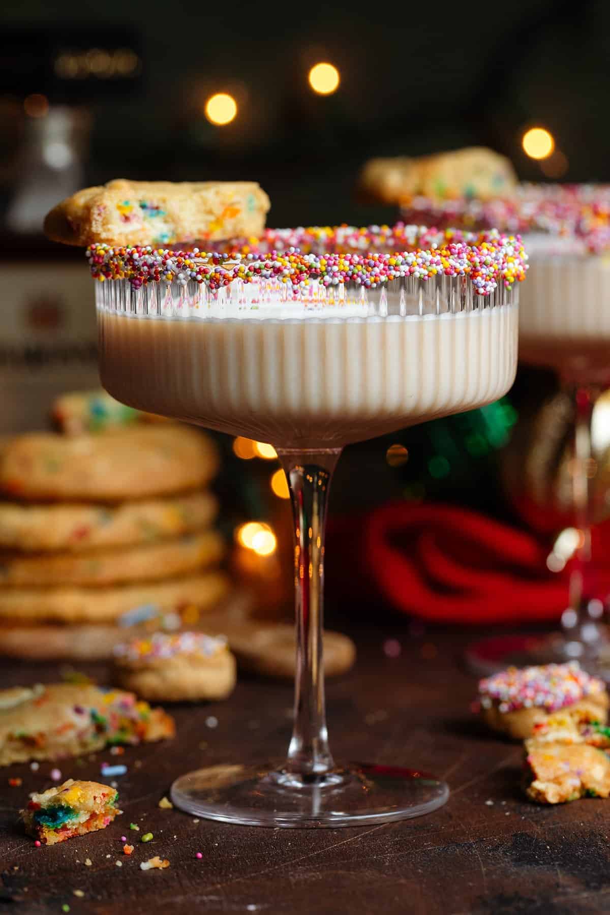 Creamy sugar cookie martini in a coupe glass with a colorful sprinkle-rimmed edge, surrounded by frosted sugar cookies with sprinkles and warm bokeh lights in the background.