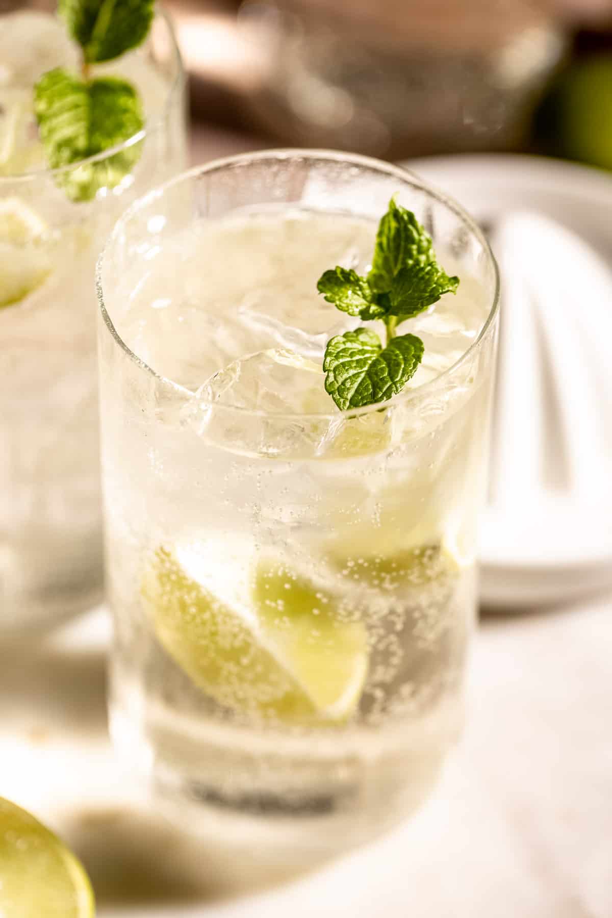 A tequila and Sprite cocktail in a tall glass filled with ice, garnished with fresh mint leaves and lime wedges, with a second drink in the background.