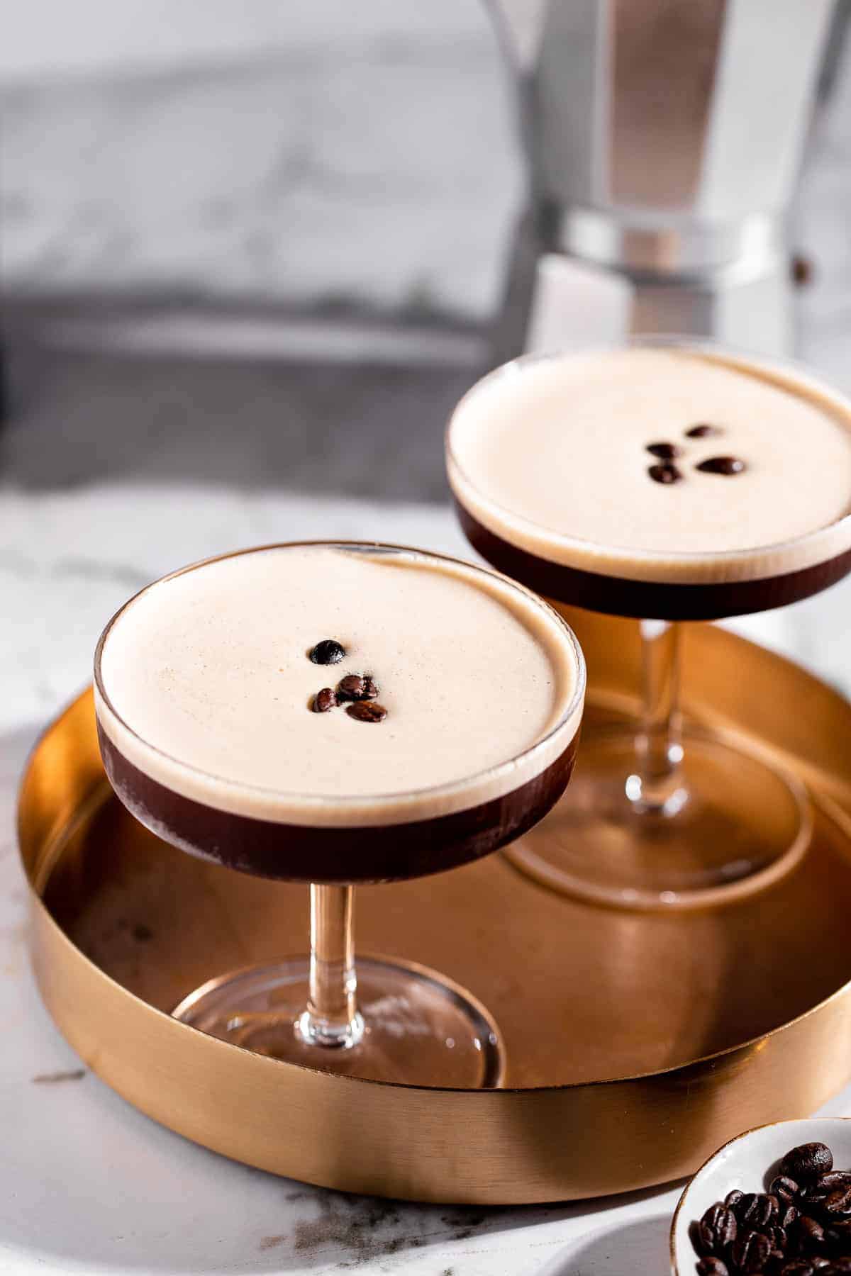 Two tequila espresso martinis in coupe glasses with silky foam tops garnished with coffee beans, served on a gold tray with a moka pot in the background.