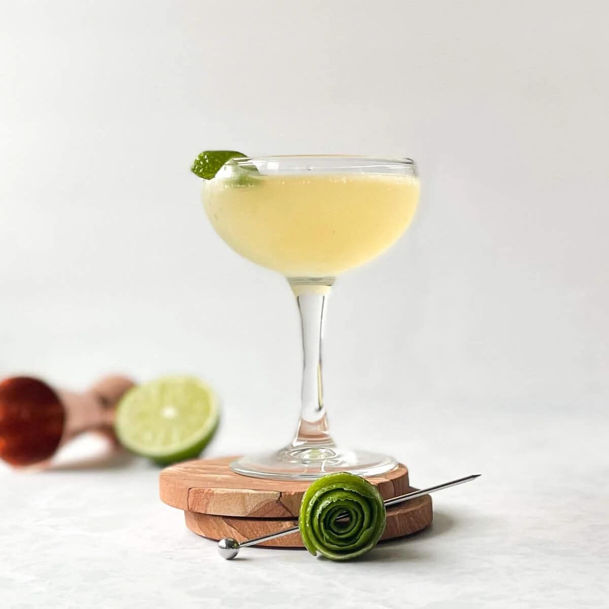 A tequila gimlet in a coupe glass garnished with a lime wheel, placed on wooden coasters with a decorative lime rose garnish and cocktail pick in the foreground.