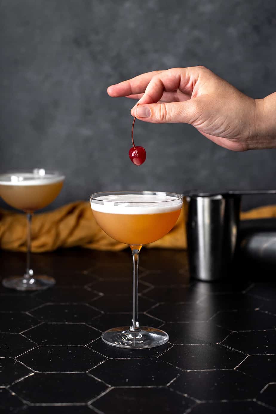 A hand placing a cherry garnish into a tequila sour cocktail served in a coupe glass with a frothy white egg white foam top, with a second cocktail and bar tools in the background.