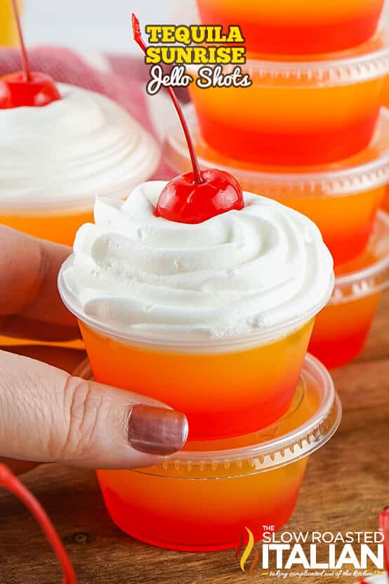 A tequila sunrise jello shot in a clear plastic cup with layered orange and red gradient colors, topped with whipped cream and a maraschino cherry, held in hand with more jello shots in the background.