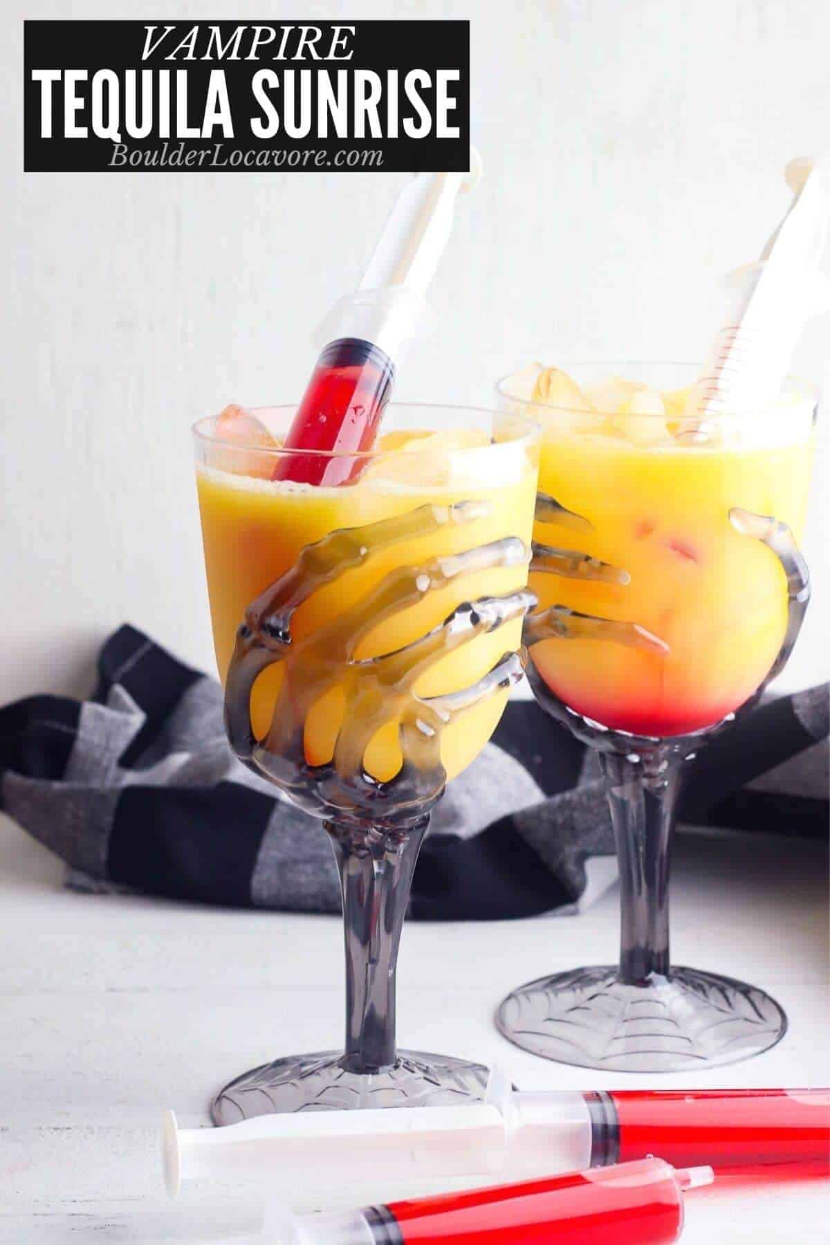 Vampire tequila sunrise cocktails in skeleton hand goblet glasses with layered orange to red colors, served with blood-filled syringe shooters for injecting grenadine, styled with black and white striped napkins for Halloween.