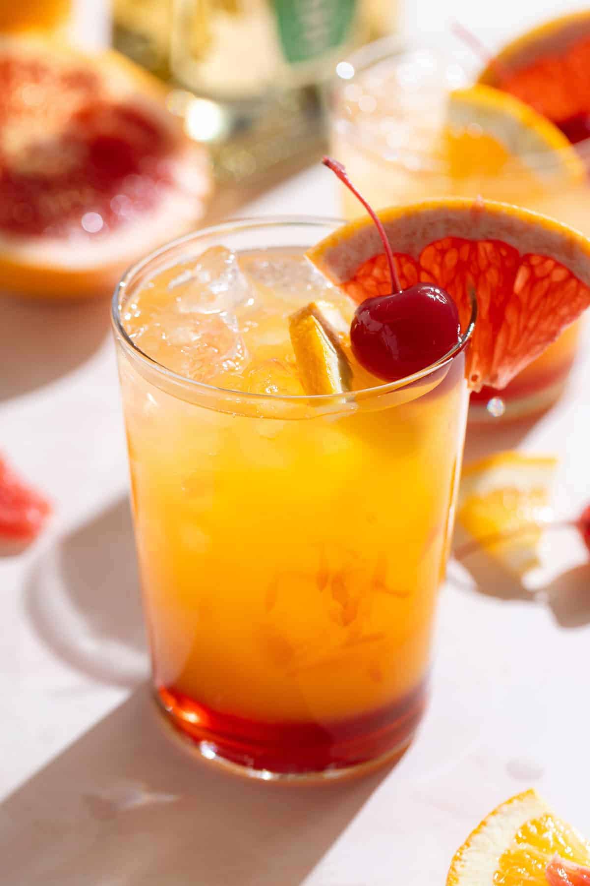A tequila sunset cocktail in a tall glass with layered yellow to red ombre colors, garnished with an orange slice, maraschino cherry, and grapefruit wedge, with fresh citrus slices scattered around.