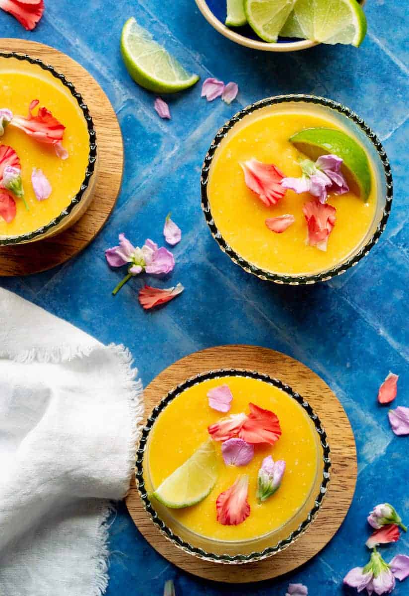 Frozen mango coconut margarita recipe in black salt-rimmed glasses garnished with lime wedges and edible flower petals on wooden coasters.