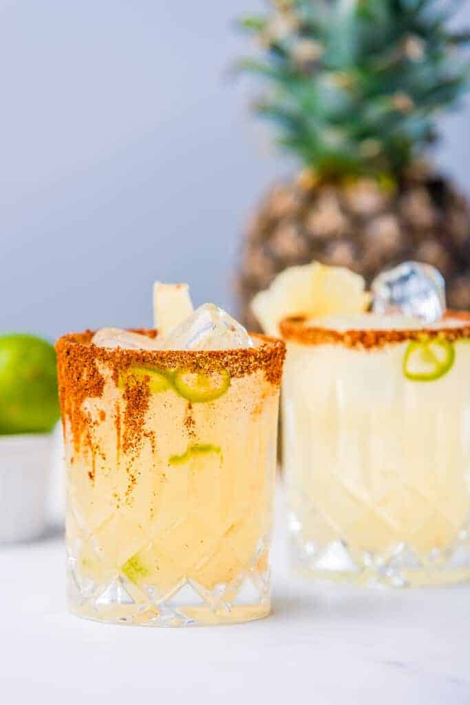 Spicy pineapple margarita recipe in crystal glasses with chili salt rim, pineapple chunks, and jalapeño slices, served with fresh pineapple.