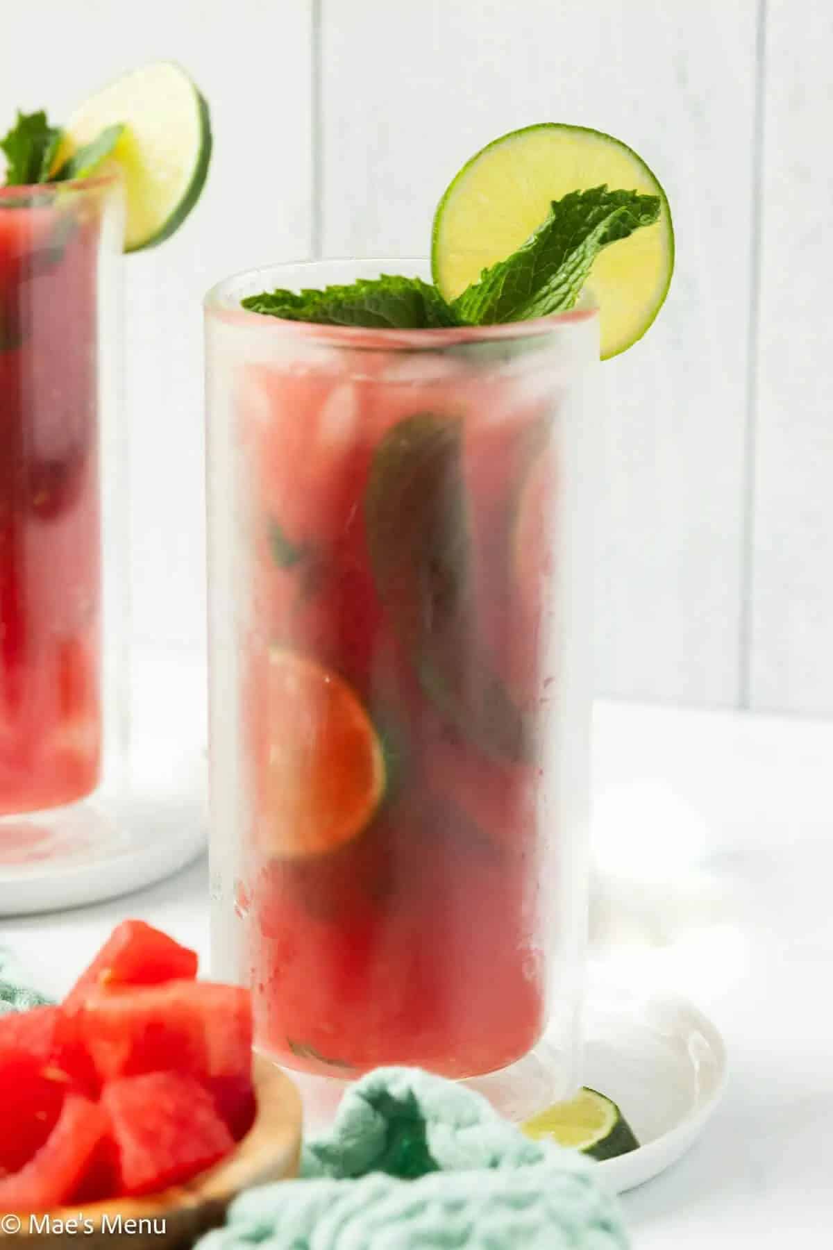 Two tall glasses of watermelon mojitos with pink liquid filled with ice, fresh watermelon chunks, and muddled mint leaves, garnished with lime wheels and mint sprigs, displayed on a white wooden surface with fresh watermelon slices and lime wedges.